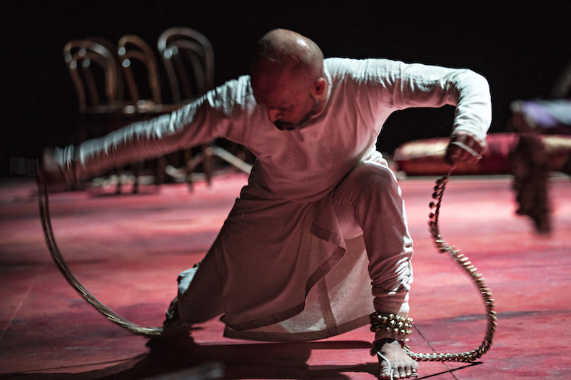 Akram Kahn crouches low to the floor. His hands hold ropes or coils that are attached to his ankles. He's wearing white.